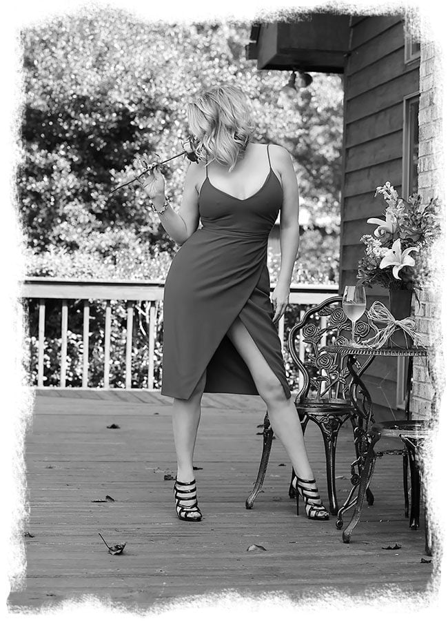 Nashville Escort Megan Love in a black and white photo, smelling a rose while wearing a sexy blue dress, standing on a wood deck with trees in the background